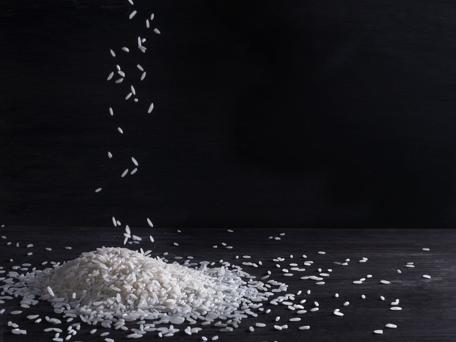 White rice grains falling into a heap on a dark wooden surface.