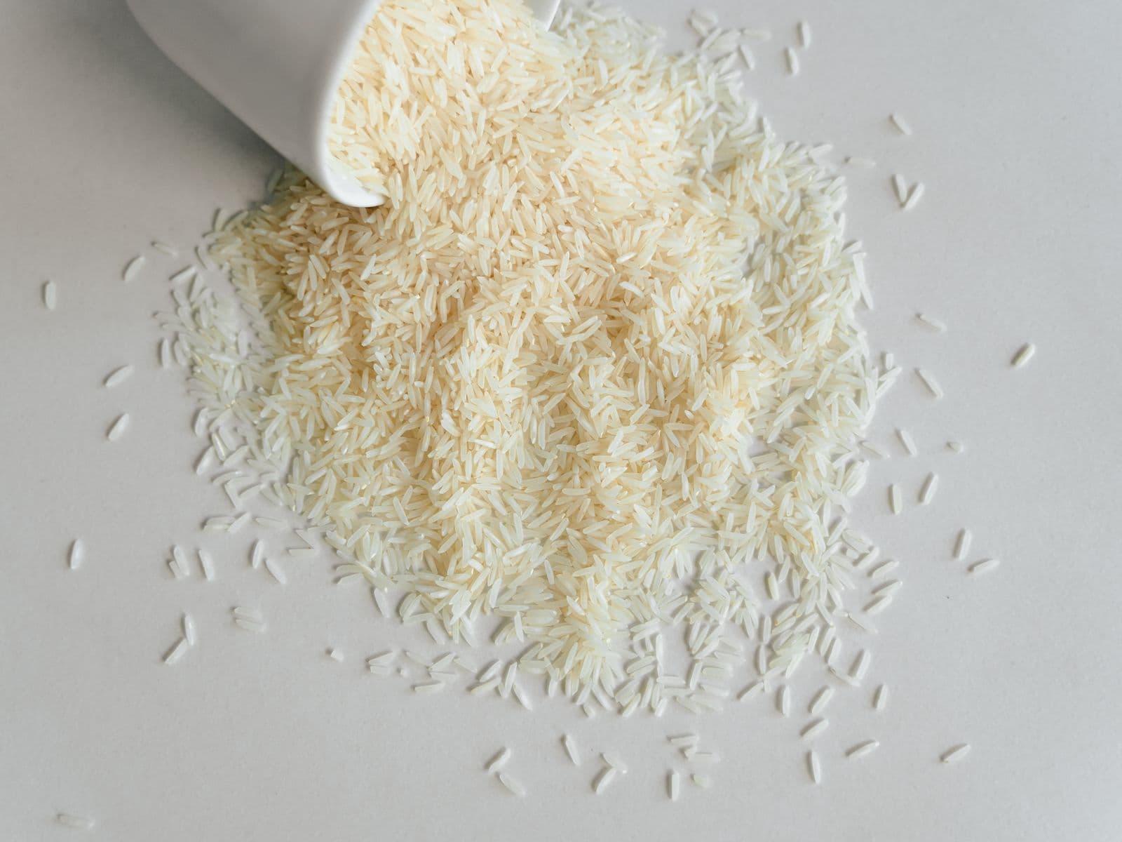 Long-grain white rice spilling from a white cup onto a pale surface.